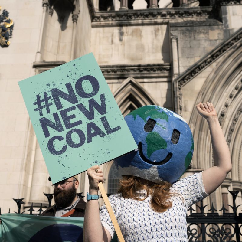 A person wearing a homemade earth-head mask holding a sign which reads "no new coal". She's stood outside the Royal Courts of Justice.