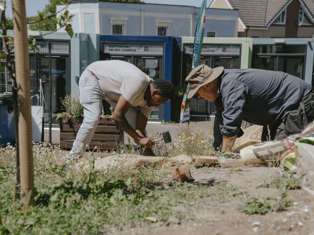 Two people crouched down gardening with shops behind them