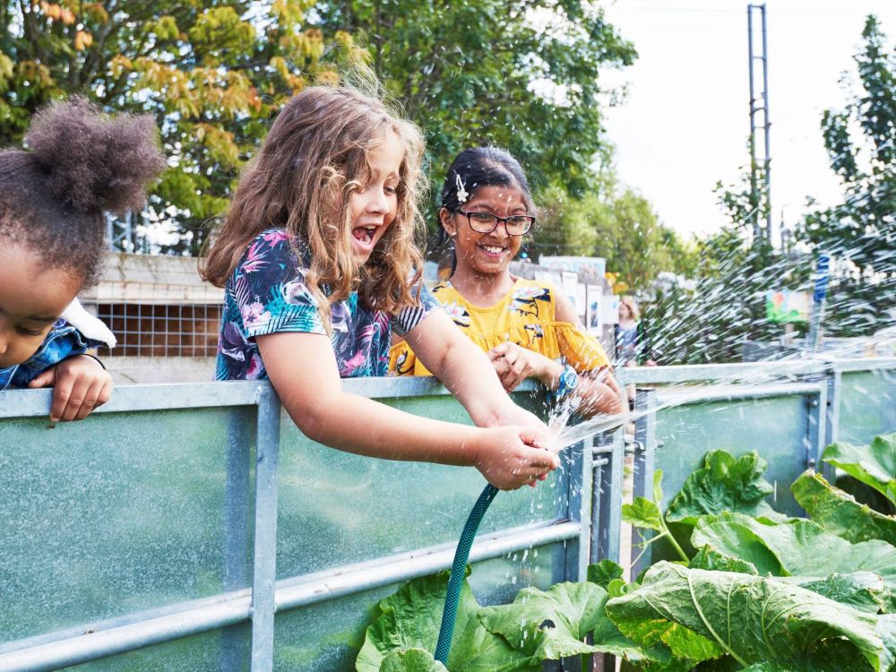 Three young girls looking into a planting area one holding a hose of splashing water