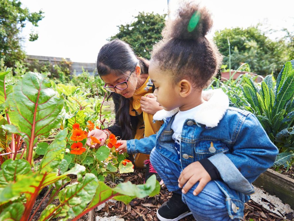 Two young girls crouched next to plants and vegetables, holding orange flowers gently in their hands.