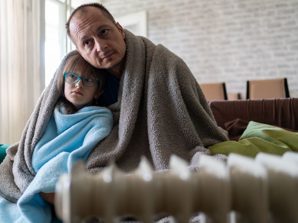 Cold father and daughter wrapped up in blankets on their sofa by a radiator