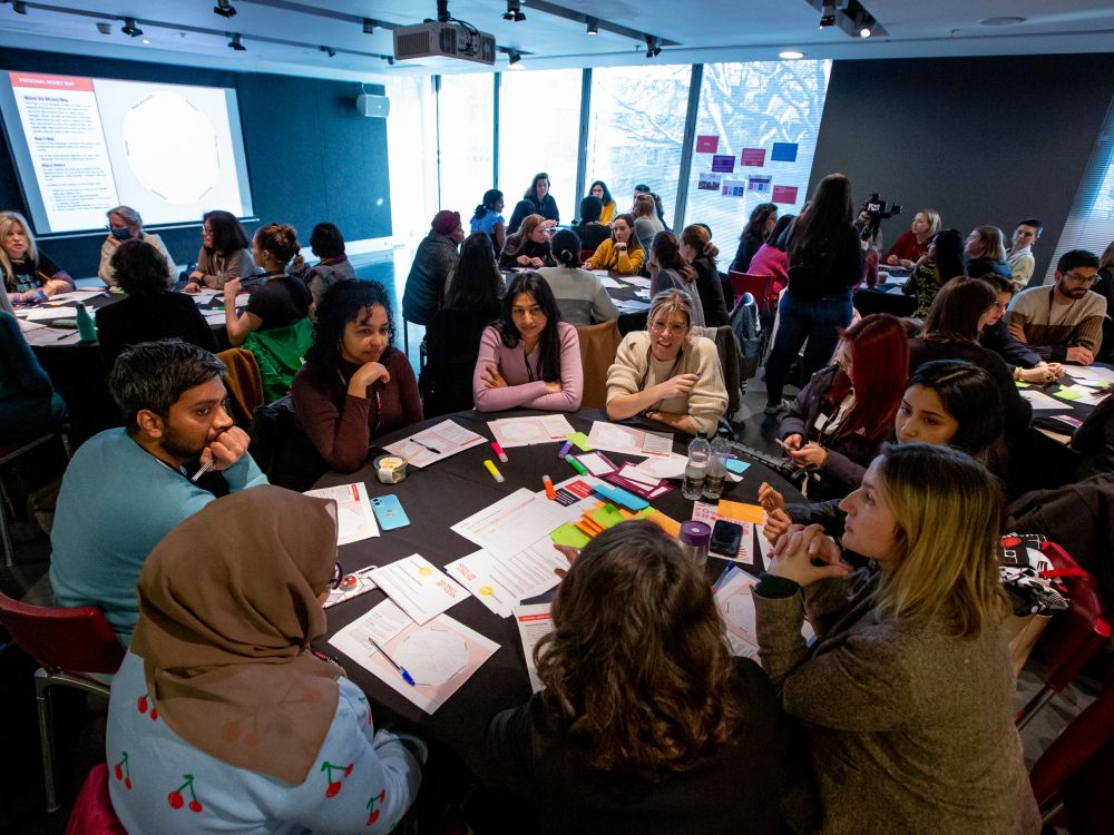 Groups of people holding discussions at round tables in a big room at a Money Movers workshop at TedX event