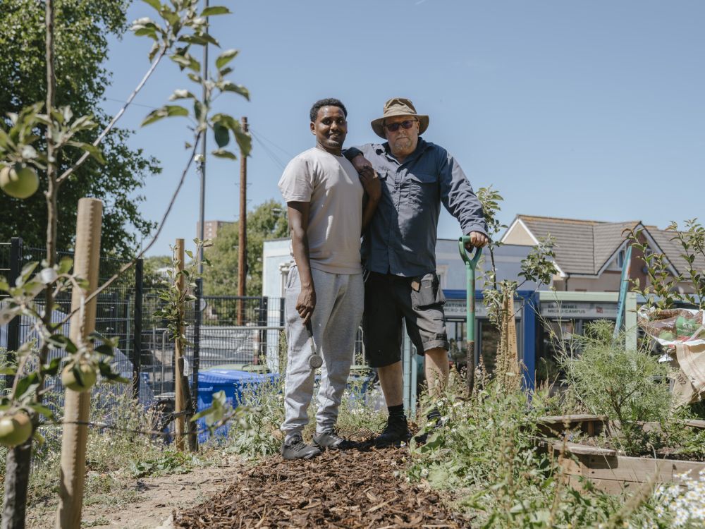 A black man and a white man lean on each other with gardening tools in an allotment near young apple trees and some shrubs with wood chips on the ground.
