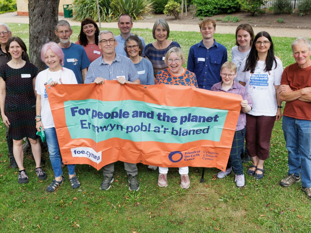 A group of people standing outside on grass with an orange banner that says 'For people and the planet' and 'Ermwyn pobl a'r blaned' and the web address  foe.cymru