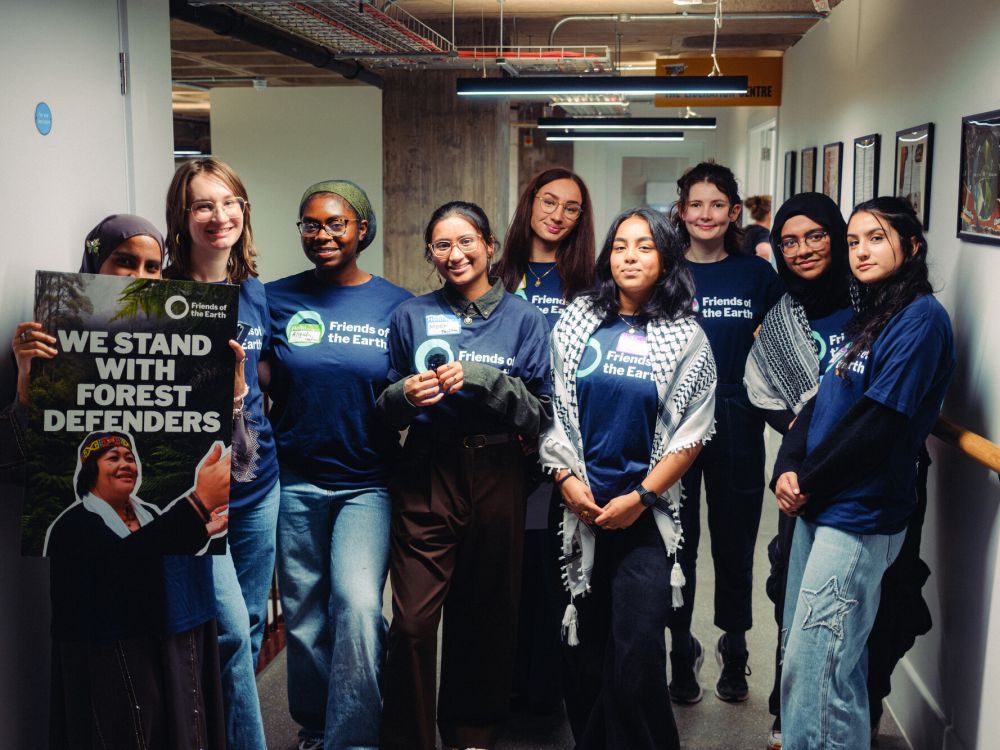 A group of young people wearing Friends of the Earth T-shirts holding a 'we stand with forest defenders' placard in a hallway.