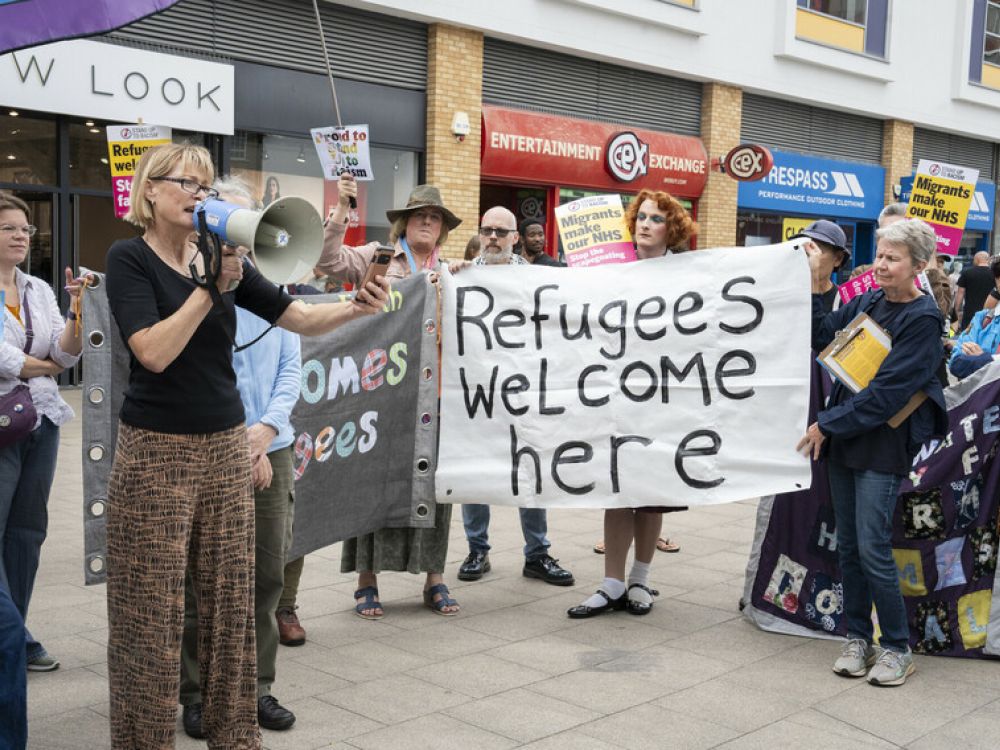 A group of people demonstrate in a high street holding banners that say "Refugees welcome here". One person is also speaking into a megaphone.